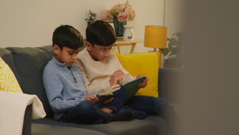 Two-Young-Boys-Sitting-On-Sofa-At-Home-Playing-Games-Or-Streaming-Onto-Digital-Tablets-11